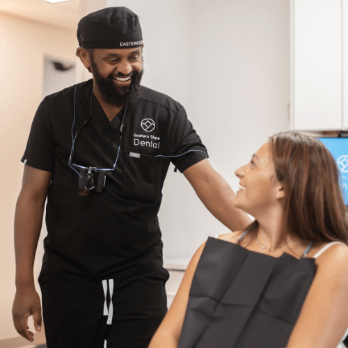 A dental professional in a black uniform stands beside a patient in a dental chair, preparing for treatment in a bright clinic setting.