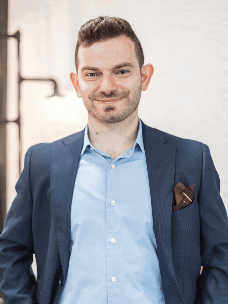 A man in a stylish navy blazer over a light blue shirt, standing confidently with his hands on his hips and a pocket square peeking out.