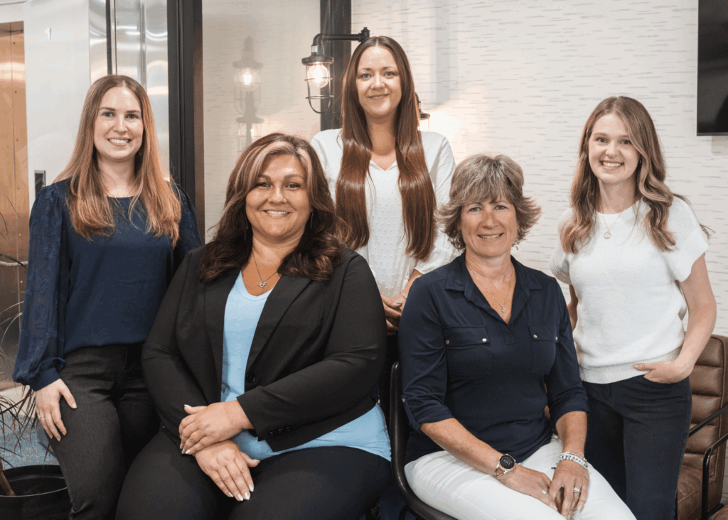 Five women pose together in a stylish, modern office space, sitting and standing, dressed in professional attire.